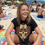 Poncho serviette à capuche motif crâne aux roses rouges, porté par une femme souriante assise sur une serviette de plage.