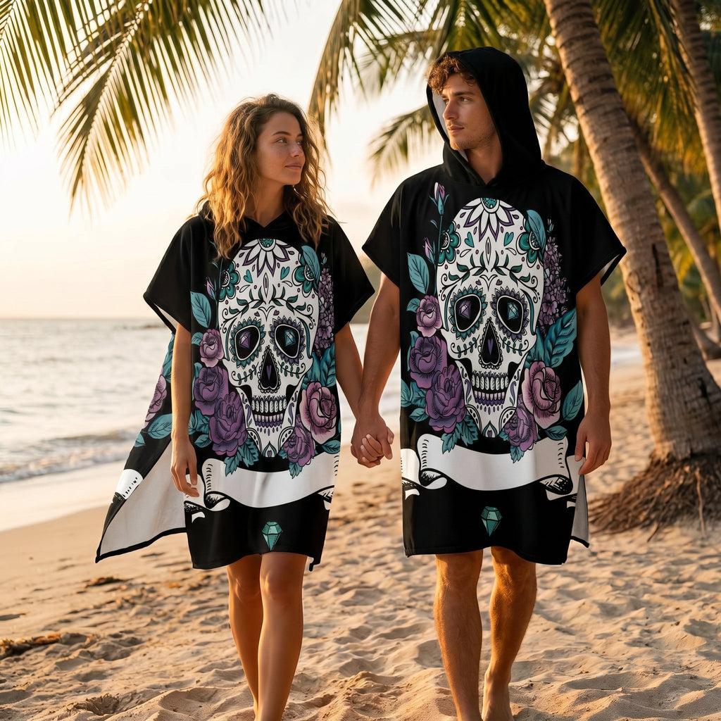 Poncho serviette a capuche motif crâne et roses violettes, porté par un couple marchant main dans la main sur une plage.