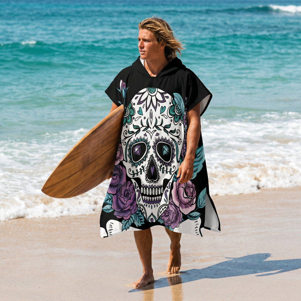 Poncho serviette a capuche motif crâne et roses violettes, porté par un homme sortant de l'eau avec sa planche de surf.