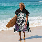 Poncho serviette a capuche motif crâne et roses violettes, porté par un homme sortant de l'eau avec sa planche de surf.