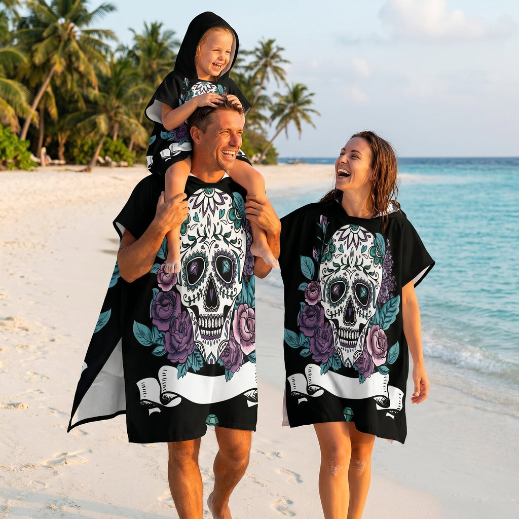Poncho serviette a capuche motif crâne et roses violettes, porté par une famille marchant sur une plage de sable fin.