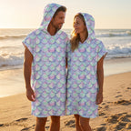 Poncho serviette a capuche motif écailles lilas, porté par un couple souriant sur une plage au coucher du soleil.