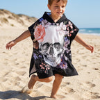 Poncho serviette a capuche motif crâne et roses pastel, porté par un garçon courant sur le sable d'une plage.