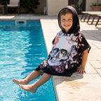 Poncho serviette a capuche motif crâne et roses pastel, porté par un garçon souriant au bord d'une piscine.