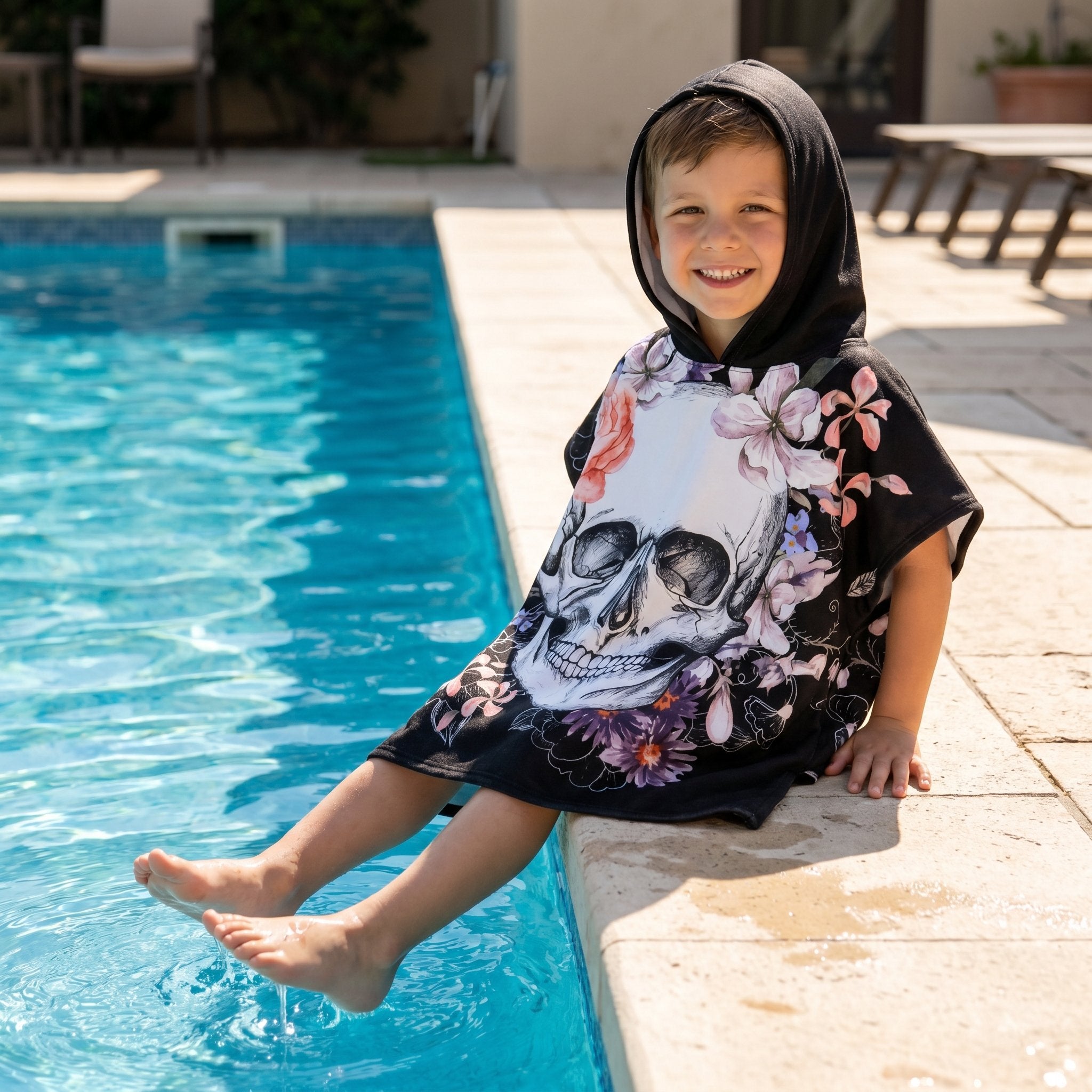 Poncho serviette a capuche motif crâne et roses pastel, porté par un garçon souriant au bord d'une piscine.