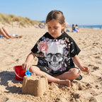 Poncho serviette a capuche motif crâne et roses pastel, porté par une fille assise sur le sable, jouant avec son seau.