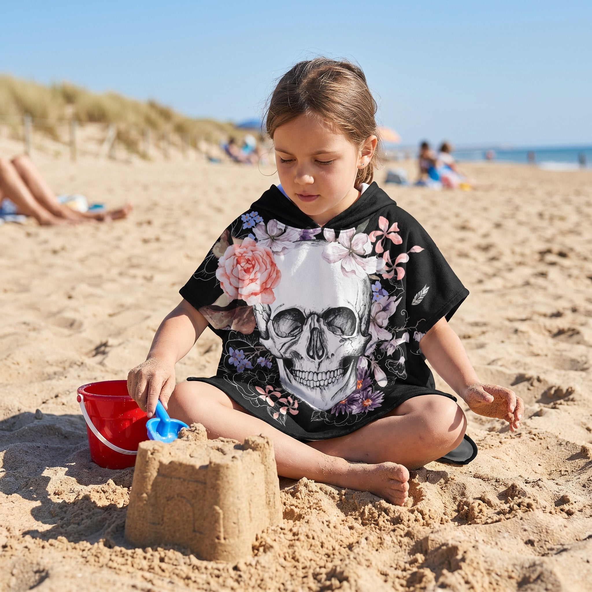 Poncho serviette a capuche motif crâne et roses pastel, porté par une fille assise sur le sable, jouant avec son seau.