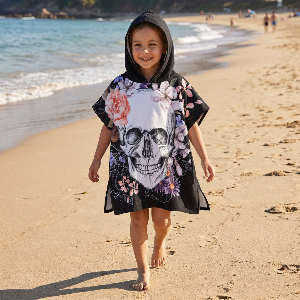 Poncho serviette a capuche motif crâne et roses pastel, porté par une fille marchant sur le sable mouillé.