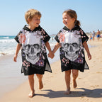 Poncho serviette a capuche motif crâne et roses pastel, portés par deux enfants joyeux qui se tiennent la main.