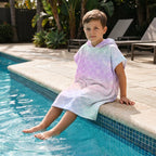 Poncho serviette a capuche motif écailles arc-en-ciel pastel, porté par un enfant assis au bord d'une piscine.