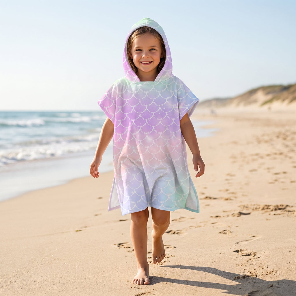 Poncho serviette a capuche motif écailles arc-en-ciel pastel, porté par une enfant marchant sur le sable de la plage.