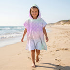 Poncho serviette a capuche motif écailles arc-en-ciel pastel, porté par une enfant marchant sur le sable de la plage.