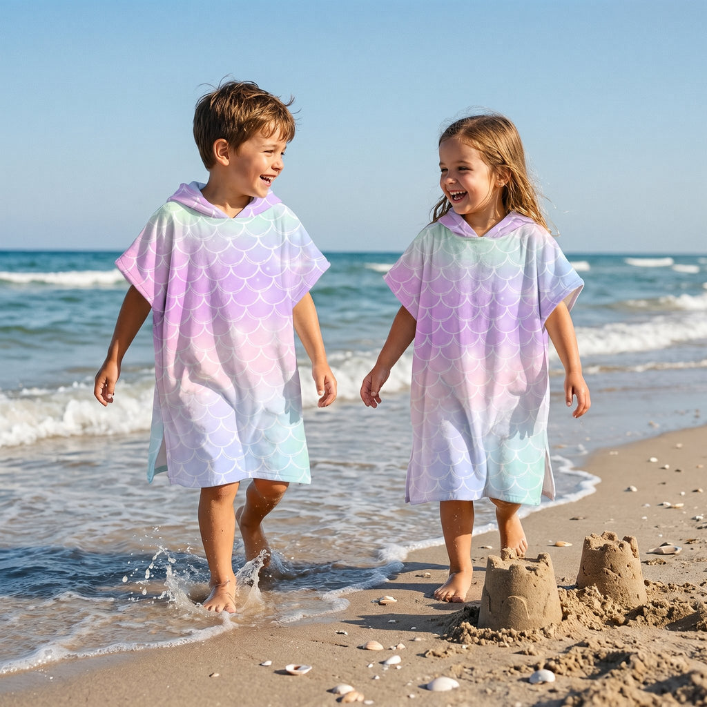 Poncho serviette a capuche motif écailles arc-en-ciel pastel, porté par deux enfants marchant au bord de l'eau.