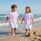 Poncho serviette a capuche motif écailles arc-en-ciel pastel, porté par deux enfants marchant au bord de l'eau.