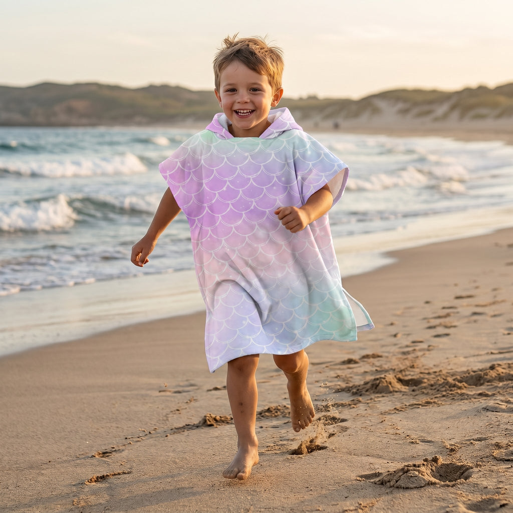 Poncho serviette a capuche motif écailles arc-en-ciel pastel, porté par un enfant courant sur le sable de la plage.