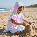 Poncho serviette a capuche motif écailles arc-en-ciel pastel, porté par une enfant construisant un château de sable.