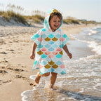 Poncho serviette a capuche motif écailles menthe et corail, porté par un garçon rieur courant dans les vagues sur la plage.