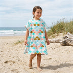 Poncho serviette a capuche motif écailles menthe et corail, porté par une fille souriante, debout sur le sable de la plage.