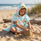 Poncho serviette a capuche motif écailles menthe et corail, porté par une fille assise, construisant un château de sable.