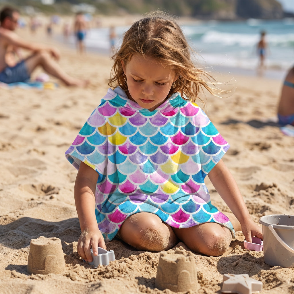 Poncho serviette à capuche motif écailles multicolores, porté par une enfant assise jouant dans le sable.