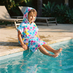 Poncho serviette à capuche motif écailles multicolores, porté par un enfant assis au bord d'une piscine.
