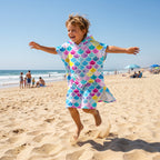 Poncho serviette à capuche motif écailles multicolores, porté par un enfant sautant joyeusement sur le sable.
