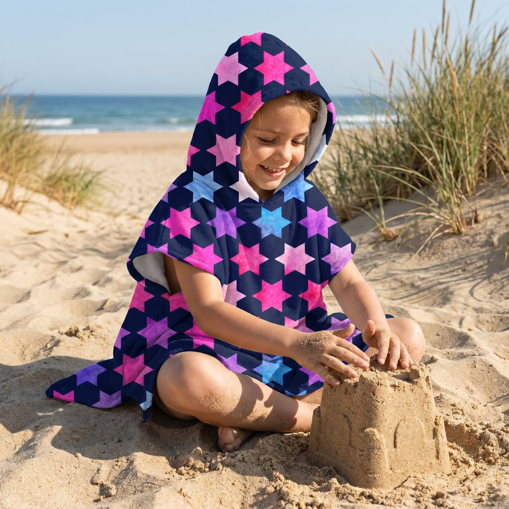 Poncho serviette à capuche enfant · Étoiles multicolores, porté par une enfant assise sur le sable