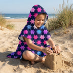 Poncho serviette à capuche enfant · Étoiles multicolores, porté par une enfant assise sur le sable