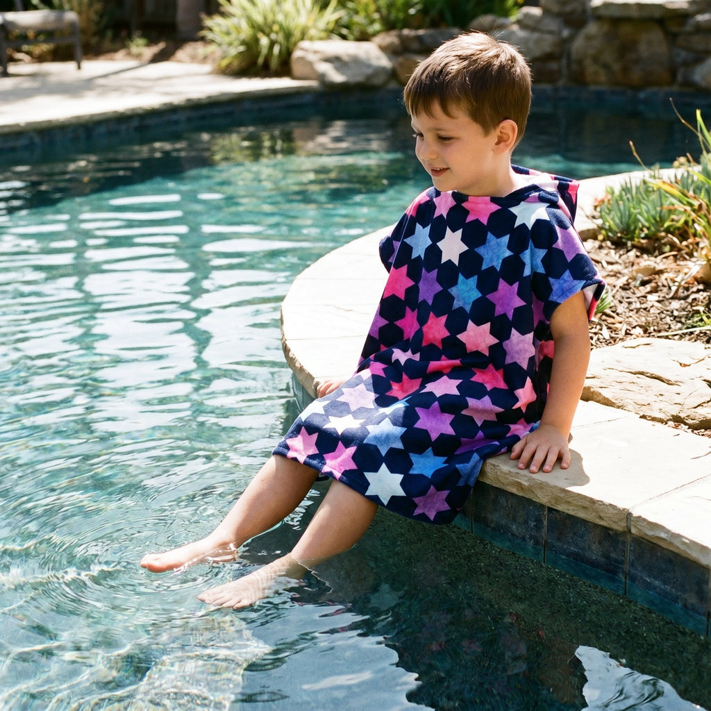 Poncho serviette à capuche enfant · Étoiles multicolores, porté par un enfant assis au bord d'une piscine