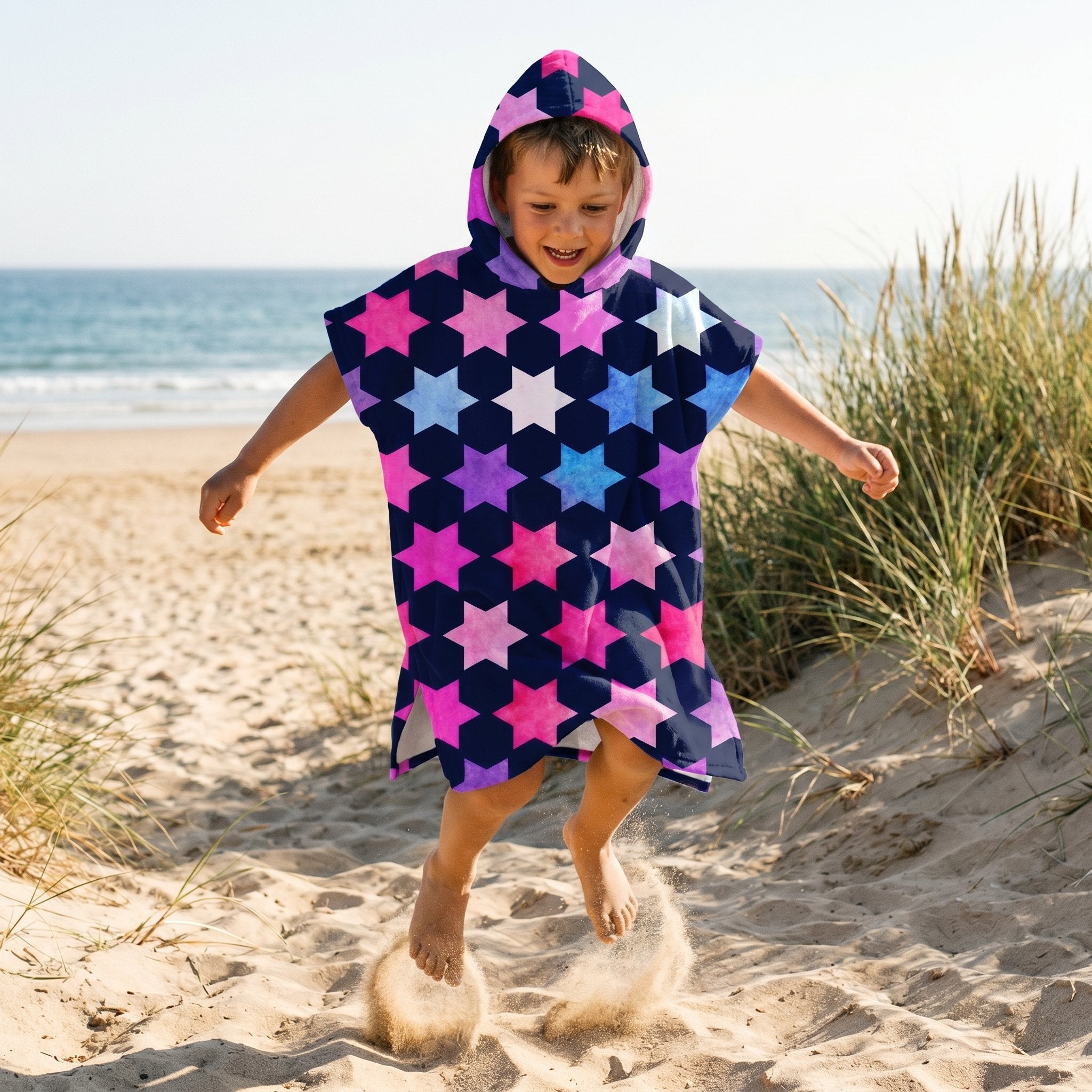 Poncho serviette à capuche enfant · Étoiles multicolores, porté par un enfant sautant sur le sable, de face