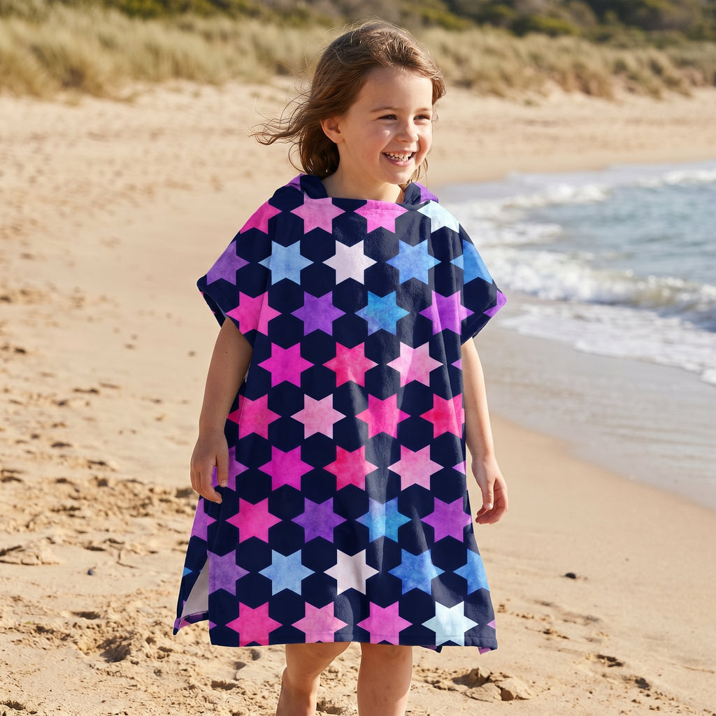 Poncho serviette à capuche enfant · Étoiles multicolores, porté par une enfant souriante, debout sur la plage