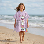 Poncho serviette a capuche motif Lama à lunettes, porté par une fille marchant sur le sable fin de la plage.