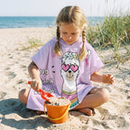 Poncho serviette a capuche motif Lama à lunettes, porté par une fille jouant dans le sable avec une pelle.