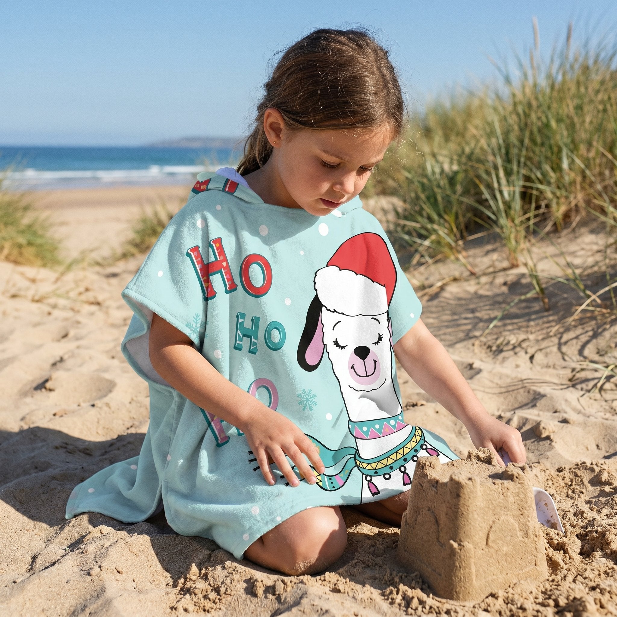 Poncho serviette à capuche motif lama bonnet de Noël, porté par une enfant assise sur le sable, jouant près de l'océan.