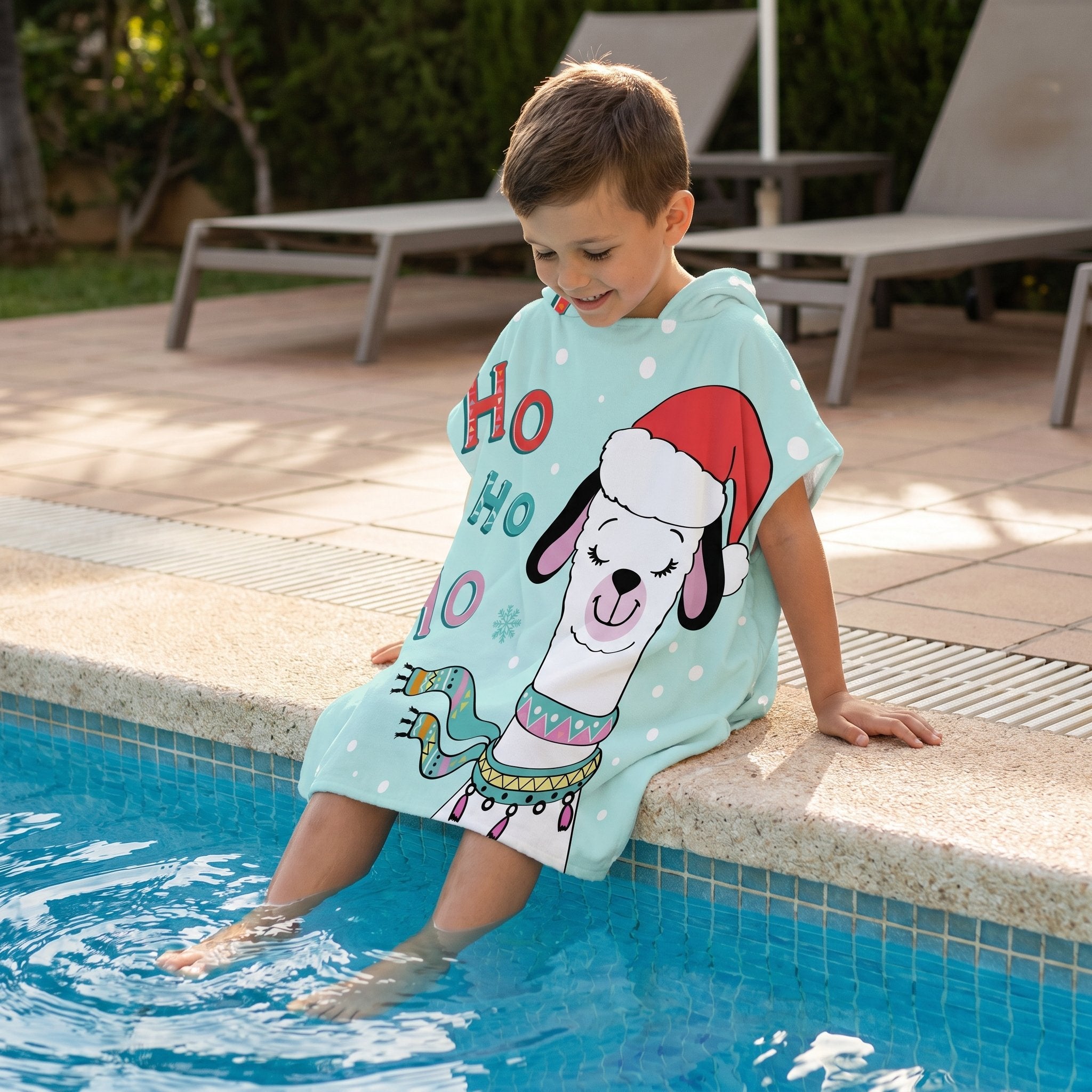 Poncho serviette à capuche motif lama bonnet de Noël, porté par un enfant assis au bord d'une piscine, les pieds dans l'eau.