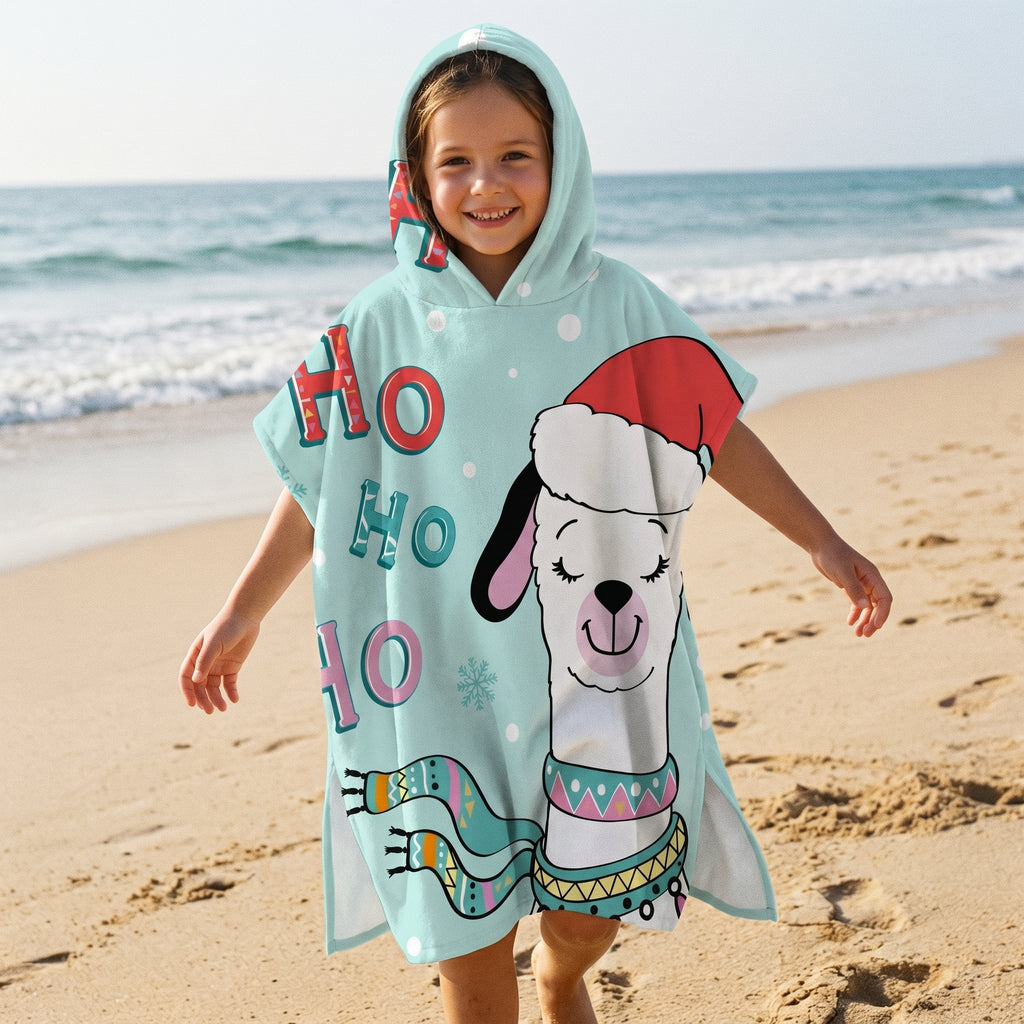Poncho serviette à capuche motif lama bonnet de Noël, porté par une enfant souriante, debout sur le sable face à l'océan.