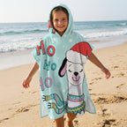 Poncho serviette à capuche motif lama bonnet de Noël, porté par une enfant souriante, debout sur le sable face à l'océan.