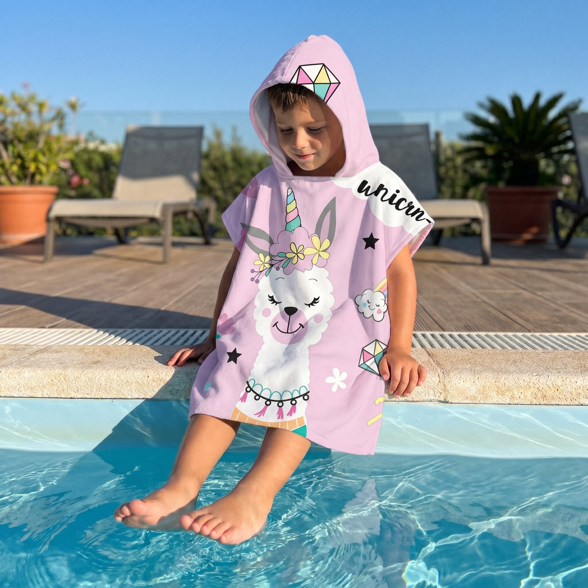 Poncho serviette a capuche motif lama licorne, porte par un enfant assis au bord d'une piscine.