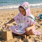 Poncho serviette a capuche motif lama licorne, porte par une enfant jouant dans le sable.