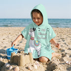 Poncho serviette à capuche motif lama menthe, porté par une enfant jouant dans le sable de la plage.