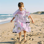 Poncho serviette a capuche motif lamas et arcs-en-ciel, porté par un enfant sautant sur la plage.