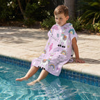 Poncho serviette a capuche motif lamas et arcs-en-ciel, porté par un enfant assis au bord de la piscine.