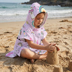 Poncho serviette a capuche motif lamas et arcs-en-ciel, porté par une enfant construisant un château de sable.