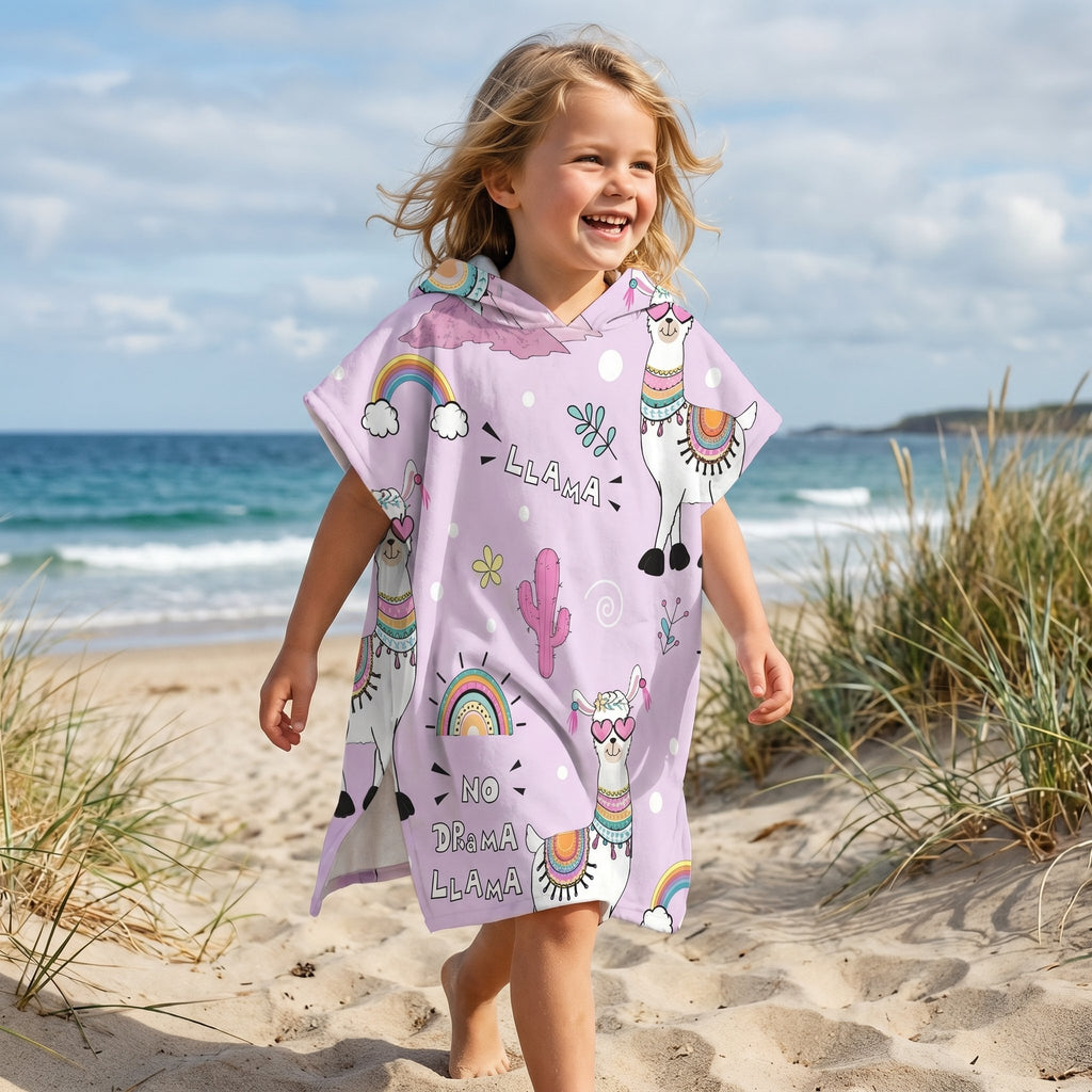 Poncho serviette a capuche motif lamas et arcs-en-ciel, porté par une enfant souriante marchant sur la plage.