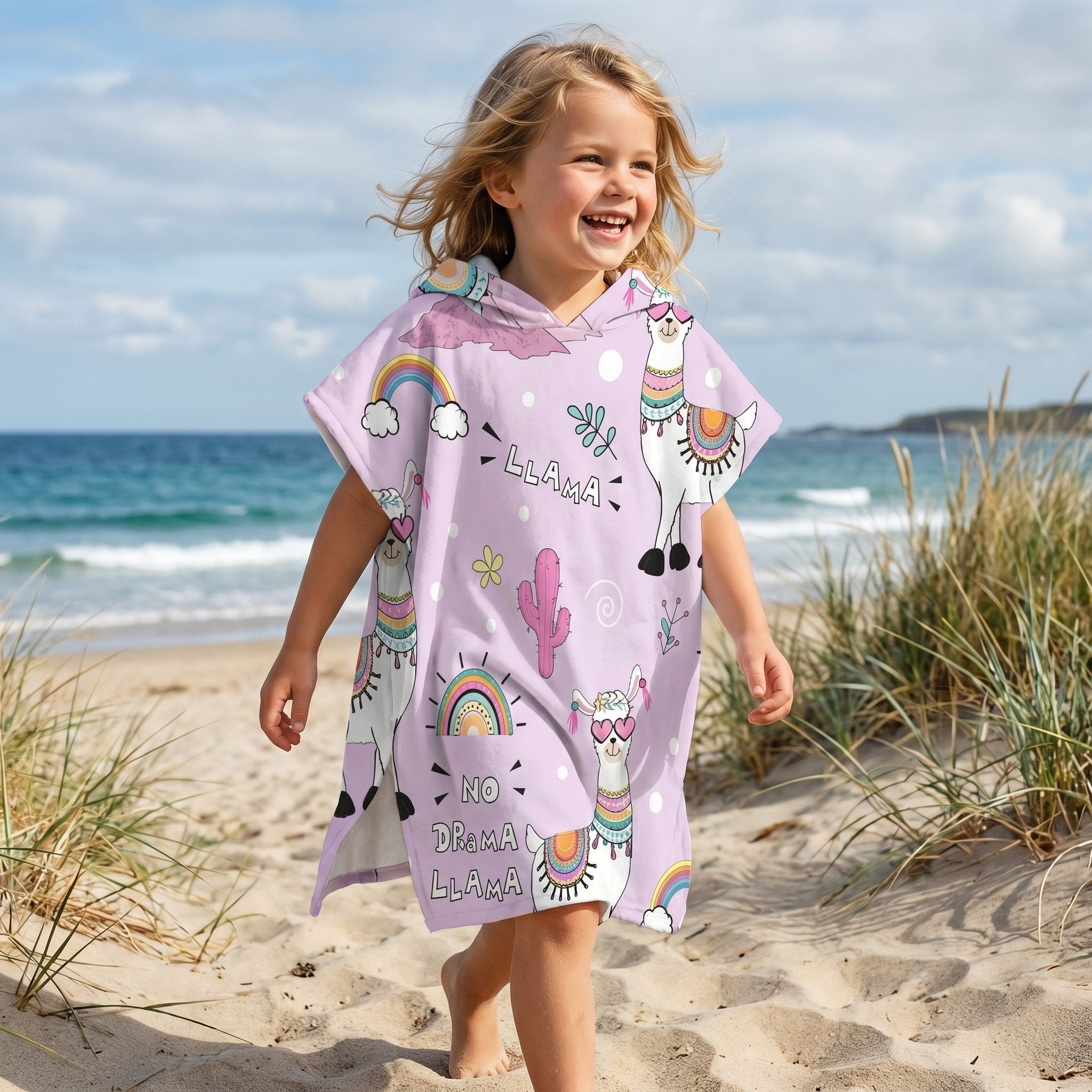 Poncho serviette a capuche motif lamas et arcs-en-ciel, porté par une enfant souriante marchant sur la plage.