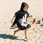 Poncho serviette a capuche motif licorne étoilée, porté par un enfant courant sur la plage.
