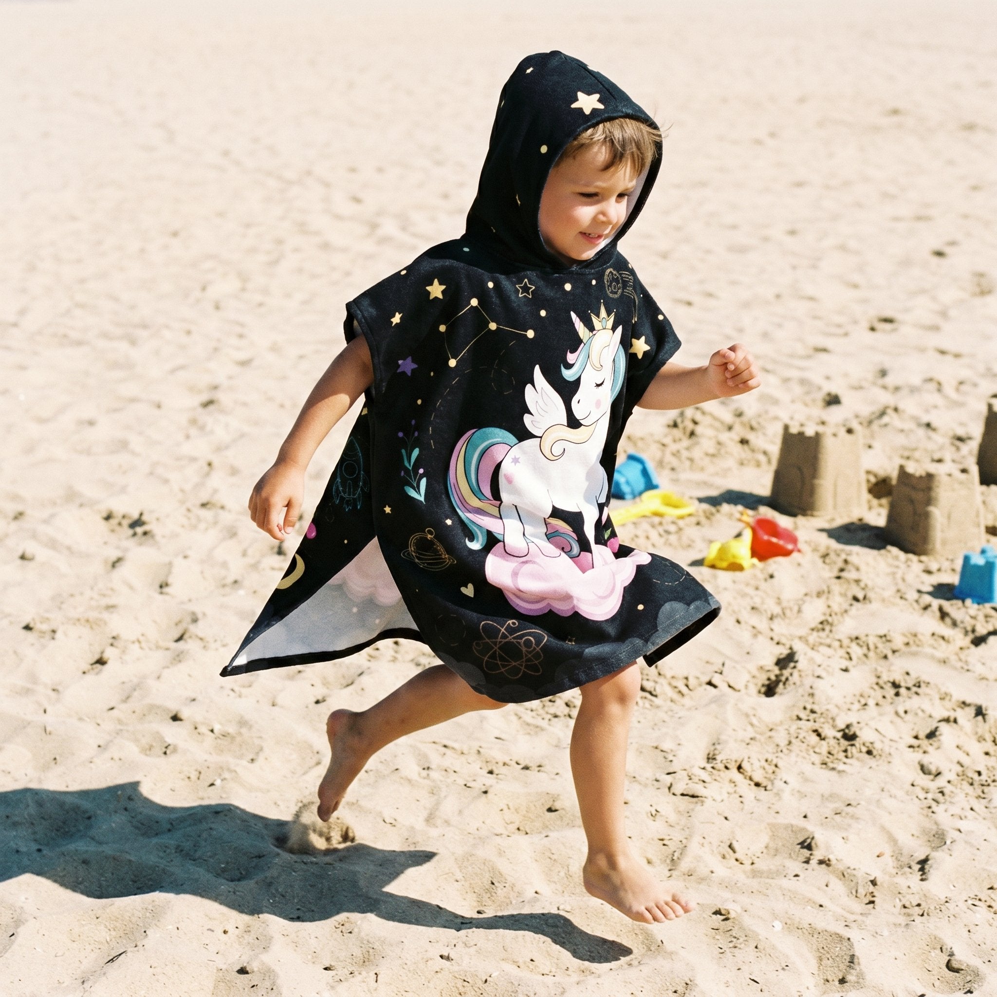 Poncho serviette a capuche motif licorne étoilée, porté par un enfant courant sur la plage.