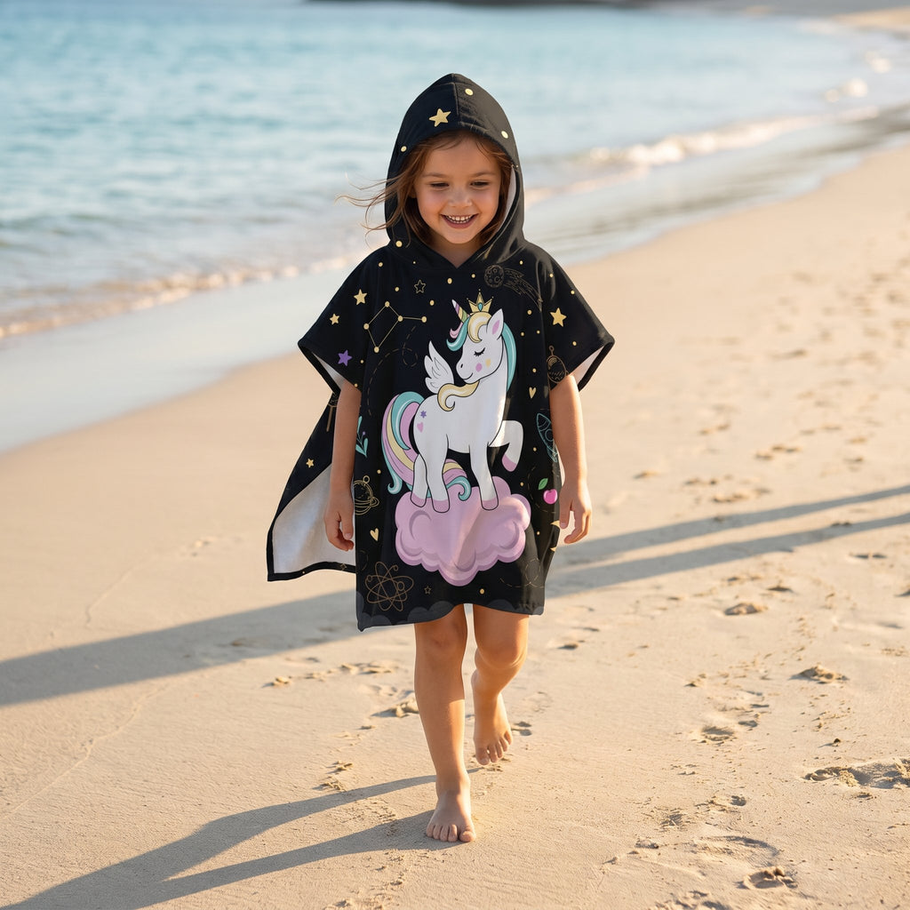 Poncho serviette a capuche motif licorne étoilée, porté par une fillette marchant sur le rivage.