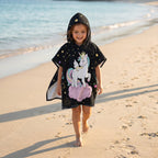 Poncho serviette a capuche motif licorne étoilée, porté par une fillette marchant sur le rivage.