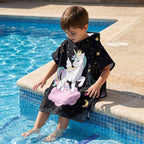 Poncho serviette a capuche motif licorne étoilée, porté par un enfant assis au bord d'une piscine.